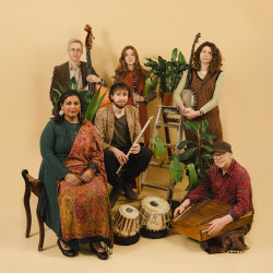 The image features the band Mishra and Deepa Shakthi sitting and standing in an ensemble holding thier instruments, with some potted plants, in front of a pale orange background