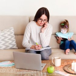 Woman taking notes while in front of the computer while young child sits behind her.