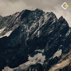 Snow-dusted rugged mountain peak under a cloudy sky, with dramatic dark rock formations. The scene conveys a sense of solitude and awe.