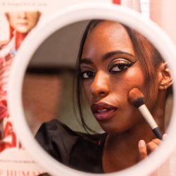Close-up portrait of a Black woman applying pink lipstick while looking into a mirror, featuring graphic black eyeliner.