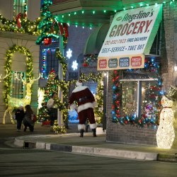 Santa Claus greets guests outside a festively decorated grocery storefront at Merry & Bright, surrounded by Christmas lights, wreaths, and illuminated holiday displays.