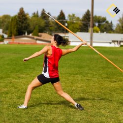 A female athlete in a red vest and black shorts is caught in mid-action, poised to throw a javelin on a grassy field. Trees and houses are in the background.