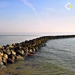 Curved rock barrier extends into calm sea under a blue sky, creating a serene, peaceful coastal scene. Cooper Education logo appears on the top right hand side.