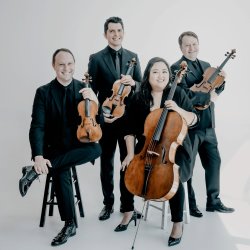 A photo of the Calidore String Quartet. The first violinist is sitting on a black stool and the second violinist is standing to his right. The cellist is sitting on a white stool in the foreground.