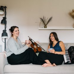 Rachel Campbell and Holli Scott are sitting with their feet up facing each other on a sofa. There is a fiddle next to Rachel and an accordion next to Holli. They are both holding a mug and laughing.