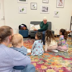 children sitting on carpet watching someone read a book