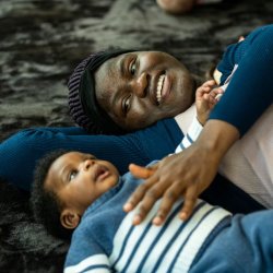 A young baby and their carer lying on their backs on a textured black rug looking up and both smiling.
