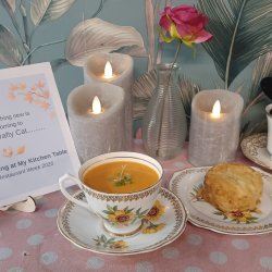 An arrangement featuring soup in a vintage tea cup, a mini cheese scone on a china plate, with candles, a little notice, and a knitted cat in a tea cup, against a background of floral wallpaper.