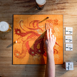 Top down photo of a hand placing a tile on the board of the game Tsuro with two pints on the table