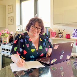 Claire dressed in a pretty black dress with colourful heart print on it sitting in her kitchen in fron of her laptop on the worktop and a sign that says a book a day keep reality away