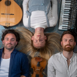 The image features Sid Goldsmith, Alex Garden and Danny Pedler lying on their backs, with Alex inverted, looking up at the camera with their instruments above their heads.