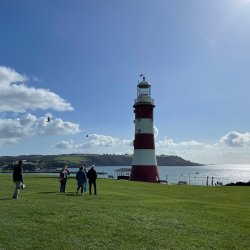 A group of people stand on green grass in front of a red and white striped lighthouse. The sea and gentle cliffs are in the background.