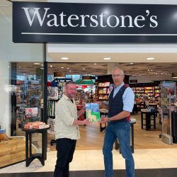 Colour Pic of Sam wearing a blue shirt with a dark blue waistcoast and blue jeans handing a copy of this book about Alpacas to Waterstone's manager Toby outside of the Waterstones Inverness Shop.