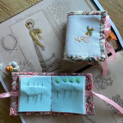 Handmade embroidered needle cases on an open book of Regency fashion illustrations, one open with needles in blue felt, tied with pink ribbon.