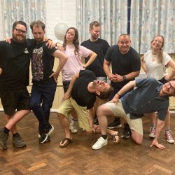 The improv team of 8, posing and laughing on a wooden floor and against light blue curtains