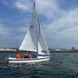 A small white sailing boat with raised sails carries a group of people wearing orange life jackets across water