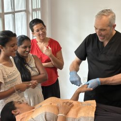 Clive Witham demonstrating a gua sha technique on the hand of a fully clothed woman who is lying on a couch. Three female students observe. They are all smiling.