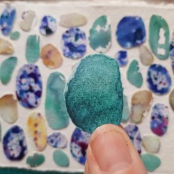 A photo of a teal coloured painted paper pebble held between finger and thumb, with a collage of pebbles in the background