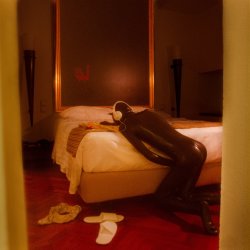 A person in a rubber suit, wearing headphones leans over a bed.