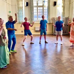 Colourful clothed singings posing in a horse shoe on a wooden floor and against magnolia walls with huge sash windows