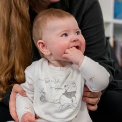 A young baby sitting up against their carer looking to the side with their hand in their mouth.