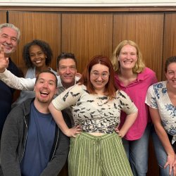 The improv team of 7 smiling against brown, wooden doors.