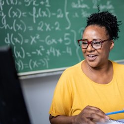 Female teacher sitting at a desk and looking at a computer in front of a blackboard. She has a pencil in her hand and is smiling.