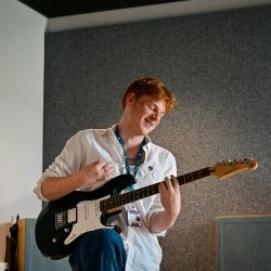 A male student wearing a white shirt is playing a black guitar infront of a grey wall.