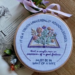 Embroidery hoop with a pastel open book and flowers design, featuring a Pride and Prejudice quote, finished with lilac ribbon on an illustrated page.