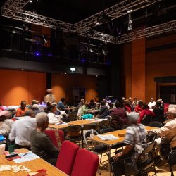 A group of people are gathered at tables at a community event in the Tramshed Theatre