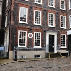 Exterior view of Dr Johnson’s House in Gough Square, London—a Georgian brick townhouse with white-framed sash windows and a dark green front door, accessed by a short flight of steps. A round commemor
