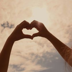 Hands in the shape of a heart against a light-filled sky