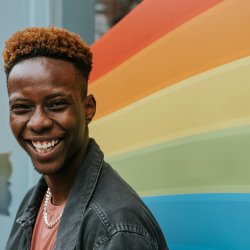 A smiling, young man stands in front of a window with a rainbow painted on it
