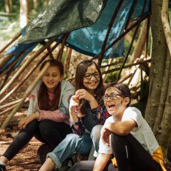 Three girls laughing in a home-made den of sticks with dirty faces.