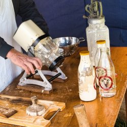 Close up of a person operating a table top butter churn with other associated items on the table beside