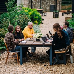 6 people working on laptops, drinking tea, deep in conversation. Sat around a wooden table, outside a flint barn, surrounded by flowers and nature.