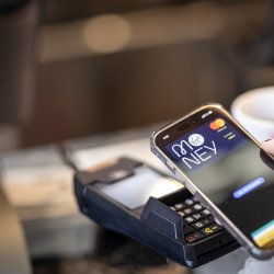 Close-up of a mobile phone displaying a Virgin Money payment screen held above a card reader on a glass counter indoors. A white cup and part of a barcode scanner are visible in the background.