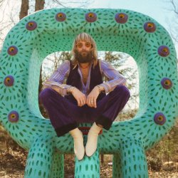 Long-haired, bearded musician (Paul McDonald) wearing sunglasses sits on a large green patterned sculptural chair in a wooded outdoor setting.