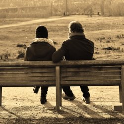 Two people sit next to each other on a park bench, their backs are facing us, they look deep in conversation