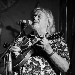 The image featuresa Phil Beer singing into a microphone and playing the mandolin in black and white