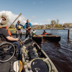 People preparing kayaks and canoes at a riverside dock, including a participant in a wheelchair wearing a life jacket, with others assisting before launching onto the water.