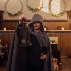 An actor wearing a cloak holds up a lantern in front of him, in his other hand is a clutch of feathers.
