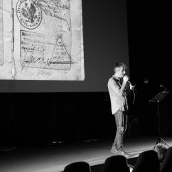Black and white image of a man speaking into a microphone, stood in front of a screen with a slide of a stamped passport page.
