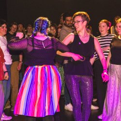 Lisa doing a right arm turn with a person wirh long blue plaited hair and a mid-length rainbow skirt in a dark room with pink lighting