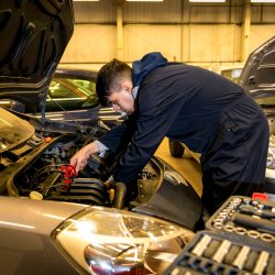 A male student in blue overalls works on a car engine