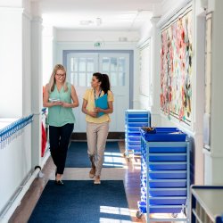Two female teachers carrying clipboards walking along a school corridor chatting and smiling.