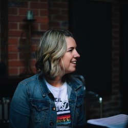 Smiling woman in a denim jacket and white T-shirt sits in profile in a rehearsal space, looking off-camera. Exposed brick walls and music stands create a warm, intimate setting.