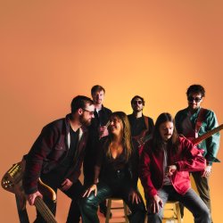Six members of The Hip Snacks pose with guitars against a warm orange studio backdrop. Two musicians sit on stools in front while the others stand behind them.