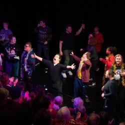 A group of performers face a seated audience with arms open wide