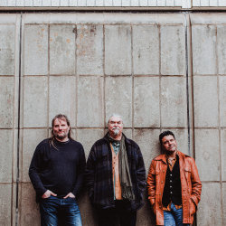 The image features Roger Flack, Peter Knight and Sacha Trochet wearing casual clothes, standing, leaning against a concrete wall